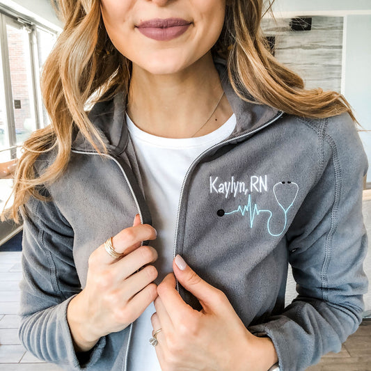 nurse wearing a gray lightweight full zip with a personalized embroidery of her name, credentials, and a heartbeat stethoscope on the left chest