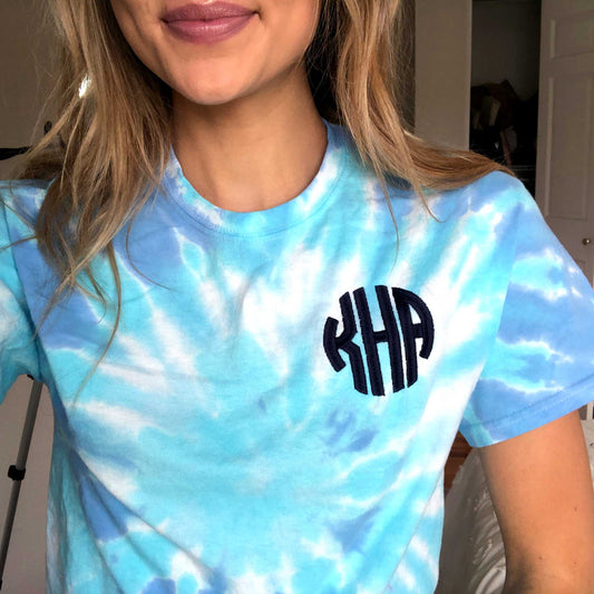 woman wearing a tie dye tee in various shades of lighter blue with a navy blue custom three letter monogram embroidery on the left chest