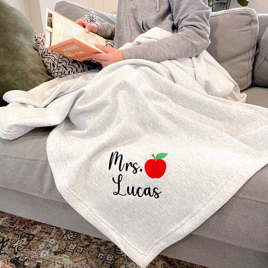 woman sitting on the couch reading with an ash  grey sweatshirt draped over her that features a name and apple embroidered design on the corner