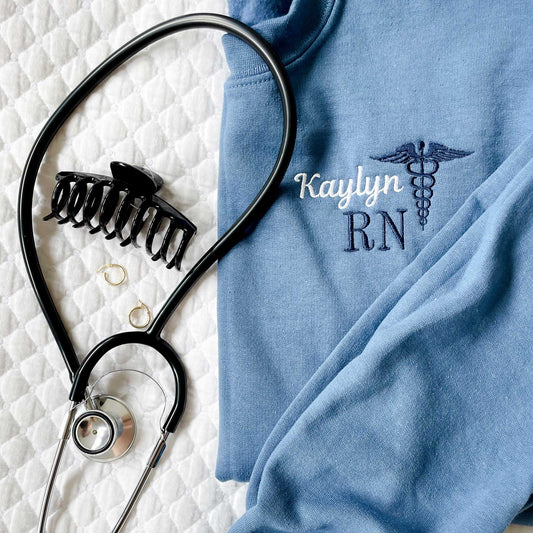 close up of a custom embroidered crewneck nurse sweatshirt next to a stethoscope, claw clip, and gold hoops