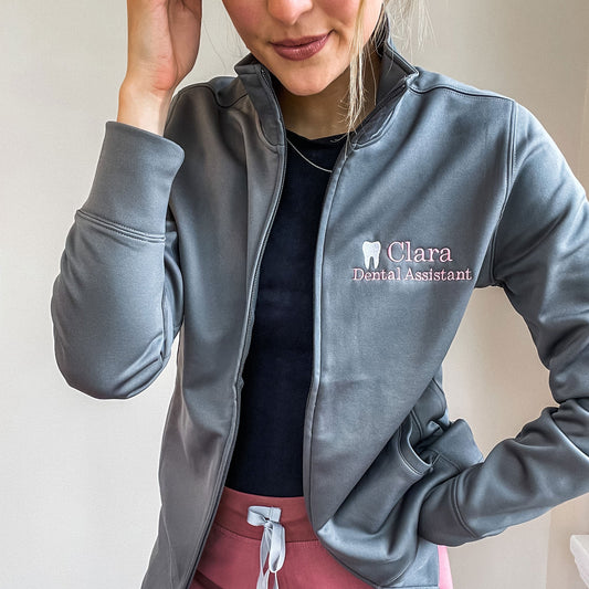 woman wearing scrub pants and a black top with a gray polyester full zip jacket over top that features a custom embroidery of her name, a mini tooth, and dental assistant underneath. 