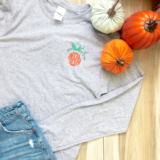 fall themed flatlay featuring a light gray long sleeved top with a custom monogram that resembles an orange pumpkin on the left chest