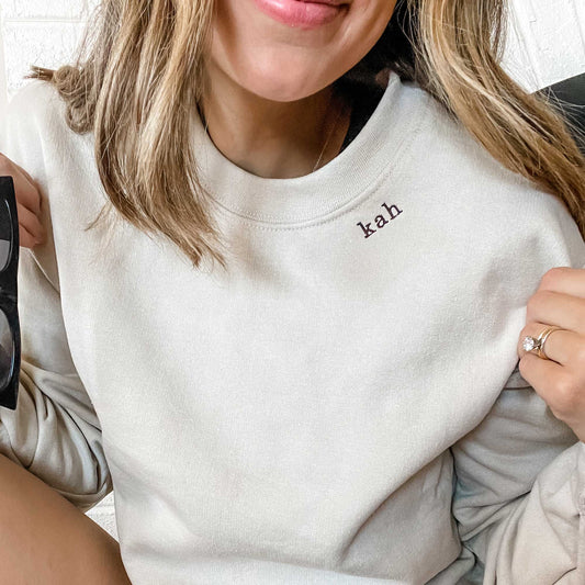 close up of a woman wearing a sand crewneck sweatshirt with embroidered mini initials in all lowercase kah on the neckline in java thread