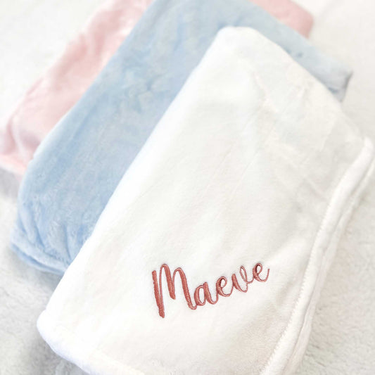 three baby blankets laying side by side in three colors with the white one showcasing a name embroidered in the corner