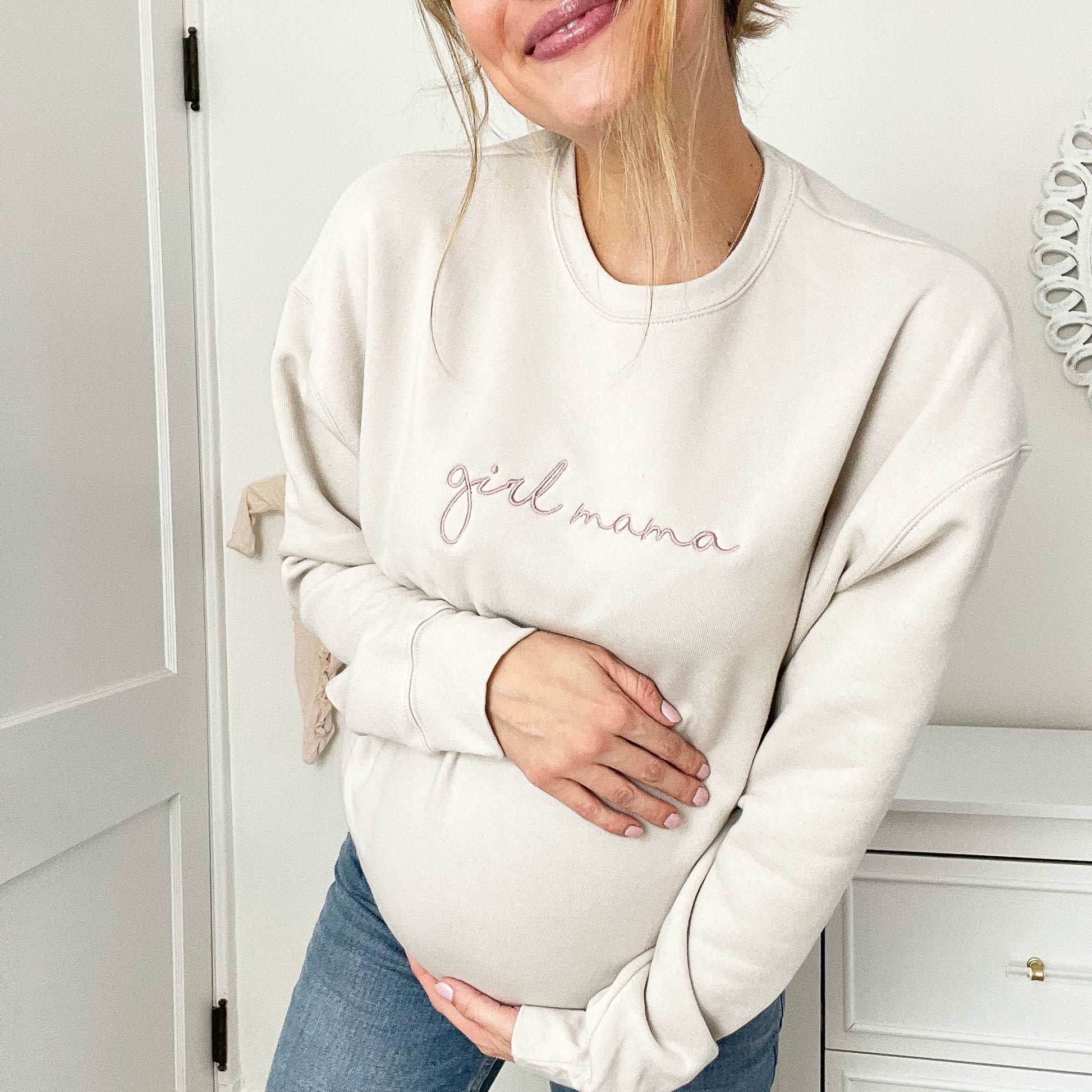 pregnant woman wearing a light beige crewneck sweatshirt with girl mama embroidered in a mauve thread across the chest