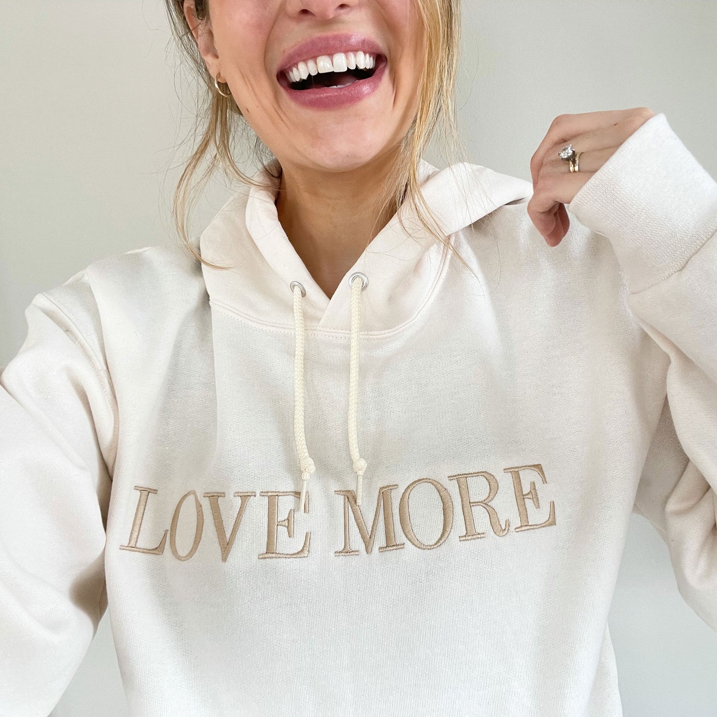 Woman wear a white hoodie that has the words Love More Embroidered across the chest in camel thread
