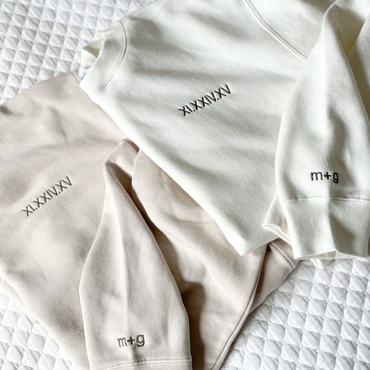two crewneck sweatshirts showcasing an embroidered roman numeral date embroidered on the left chest and matching initials on the cudd