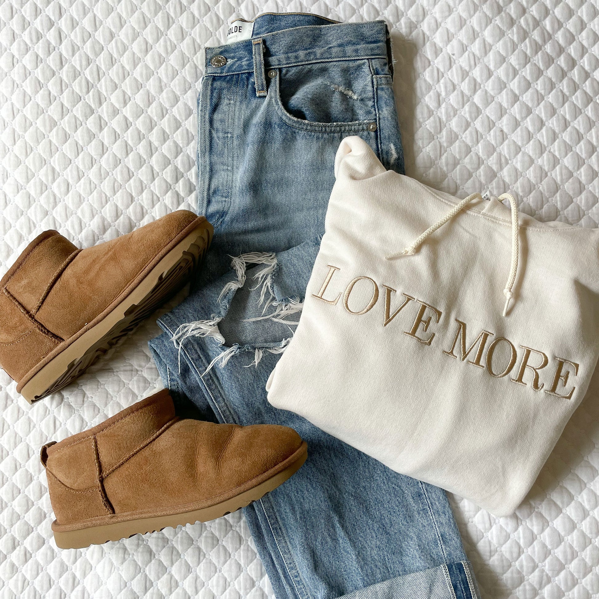 styled flat lay image of jeans and ugg boots with a white hoodie that has the text Love More embroidered across the chest
