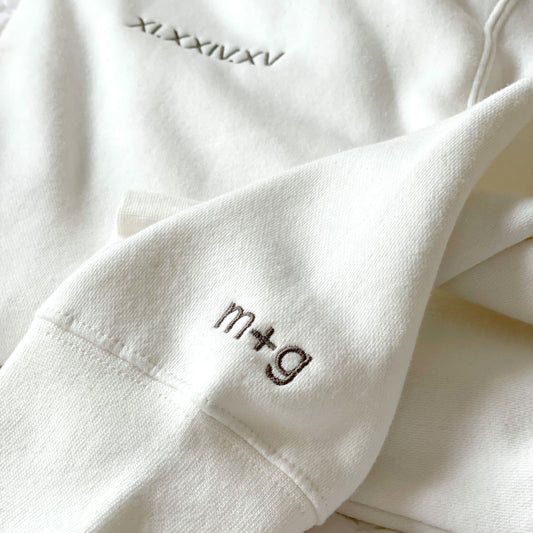 close up of a white crewneck sweatshirt showing the cuff embroidery letters m+g