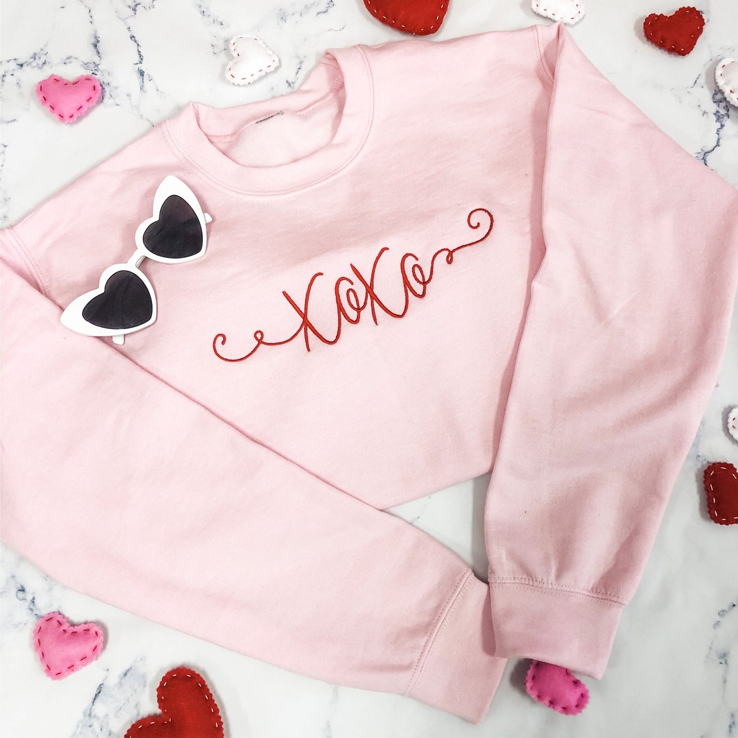flatlay valentines day themed image with felt hearts, heart framed glasses, and a light pink crewneck sweatshirt with xoxo embroidered in a script font and crimson thread across the front
