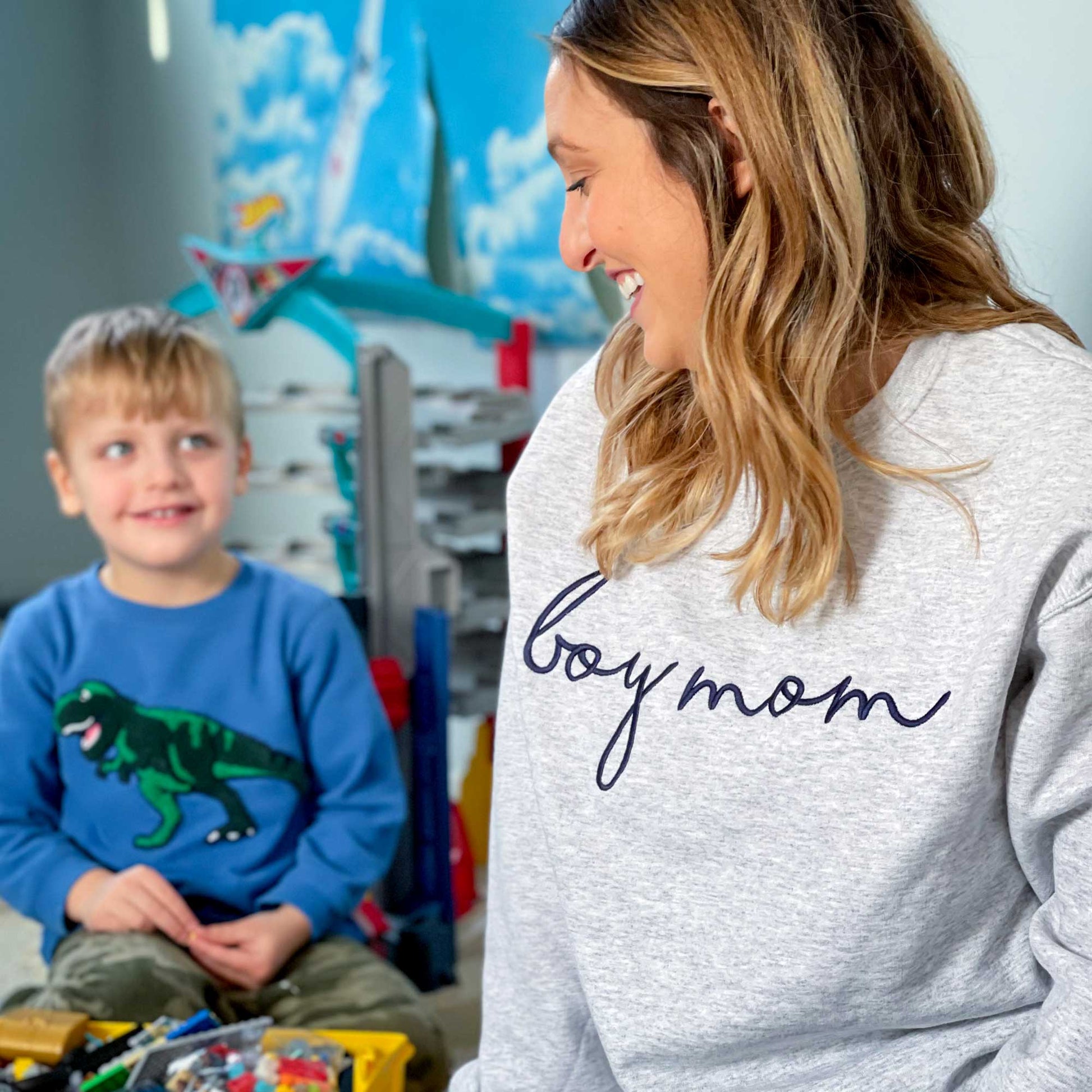 a woman sitting on the floor of her son's room wearing a light gray pullover sweatshirt with  boy mom embroidered in a cursive font and navy blue thread across the chest