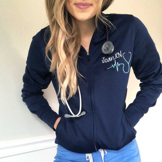 a female nurse wearing scrubs and a personalized full zip dark blue jacket with a custom embroidery on the left chest featuring her name, credentials, and a heartbeat stethoscope