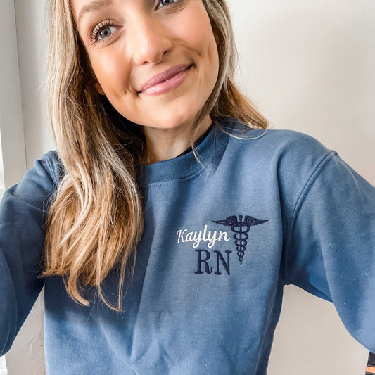 woman wearing a pullover crewneck indigo blue sweatshirt with a personalized name, credentials, and caduceus symbol embroidered in white and navy blue on the left chest