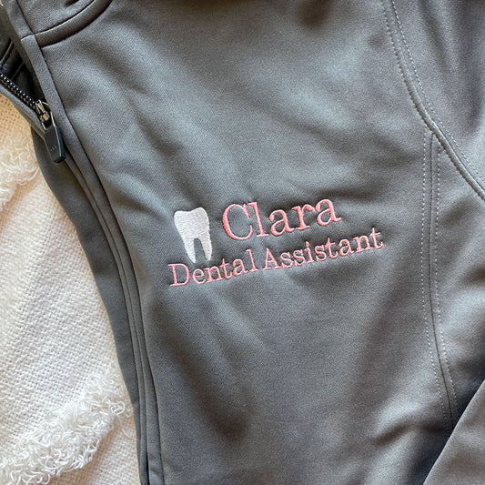 close up of a personalized dental assistant jacket  with the embroidery featuring a name, a mini white tooth, and credentials on a gray polyester jacket