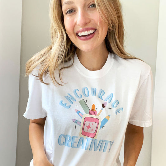 woman wearing a white comfort colors t-shirt with an 'encourage creativiey' colorful print featuring various school supplies