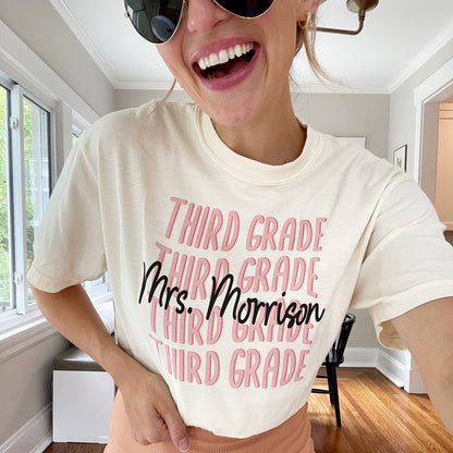 woman wearing an ivory comfort colors t-shirt with a block wvavy retro print of a grade level and teacher name centered on the shirt