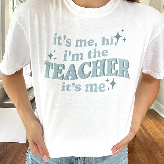 close up of a white comfort colors t-shirt with a custom "it's me, hi, i'm the teacher it's me" DTG printed deisgn across the front