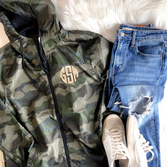 flatlay image of blue jeans, white sneakers, and a forest green camo windbreaker with black zipper and custom three letter embroidered monogram in a camel thread. 