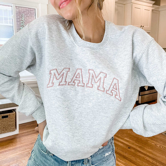 woman standing in her kitchen wearing jeans and an ash gray pullover sweatshirt with MAMA embroidered in a varsity block across the chest in a mauve thread