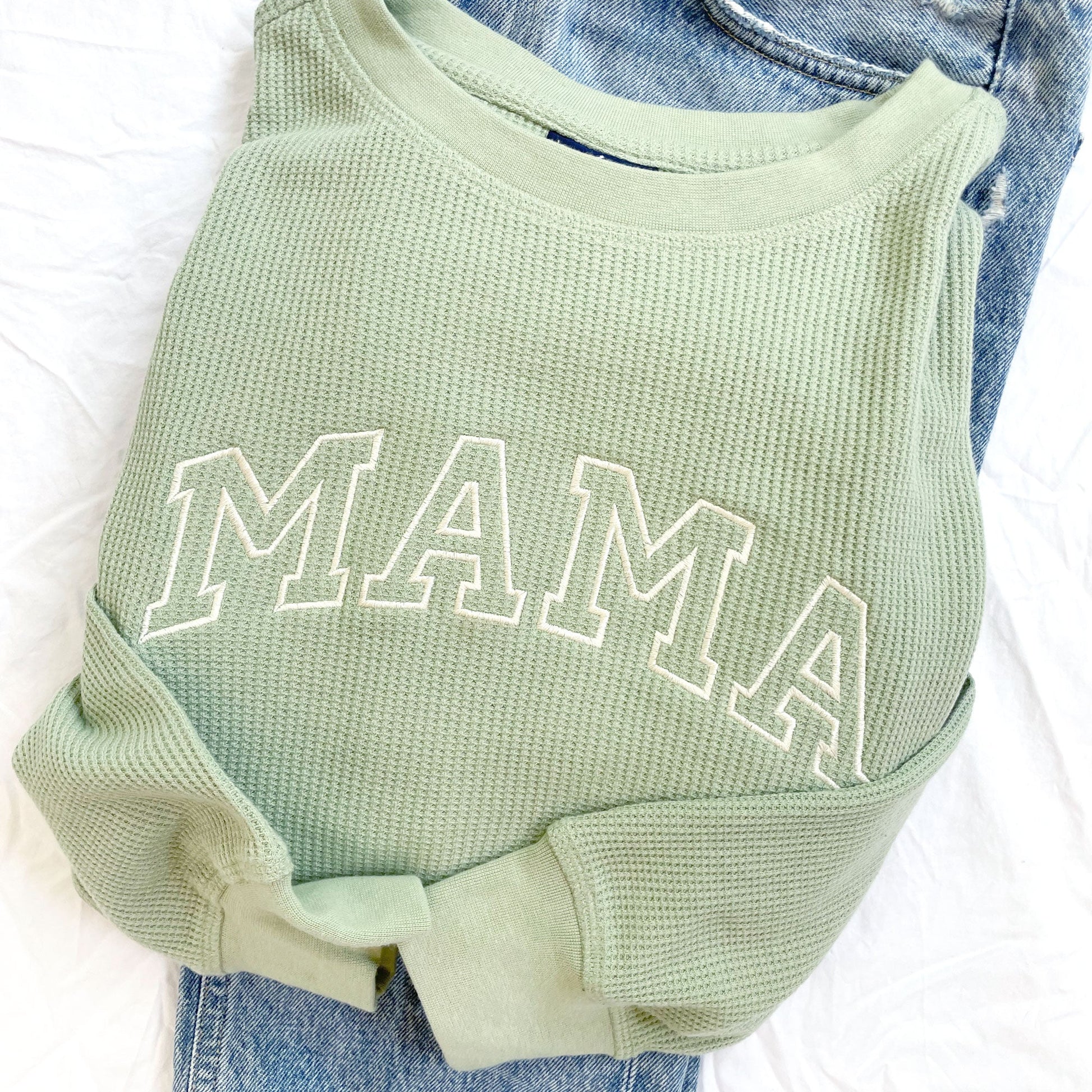 sage green crewneck with mama in embroidered outlined varsity letters across the chest folded on top of a pair of jeans