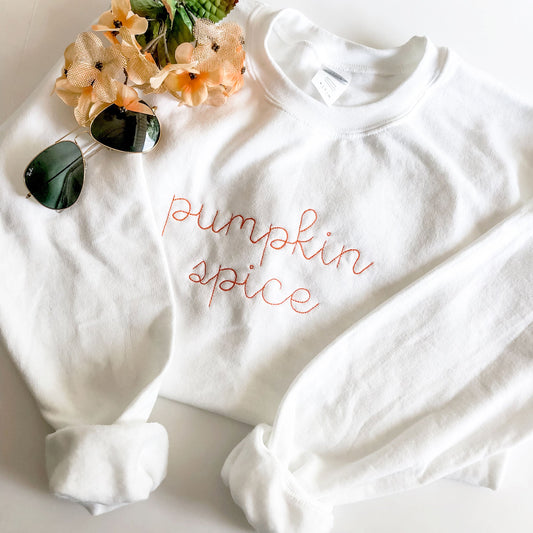 flatlay image of a white crewneck sweatshirt with pumpkin spice embroidered across the chest in a orange thread.