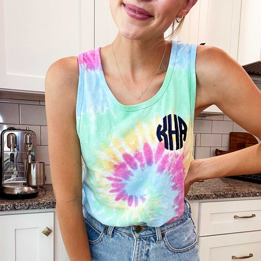 young woman wearing jean shorts and pastel spiral dyed tank top in pastel colors with a navy blue monogram on the left chest