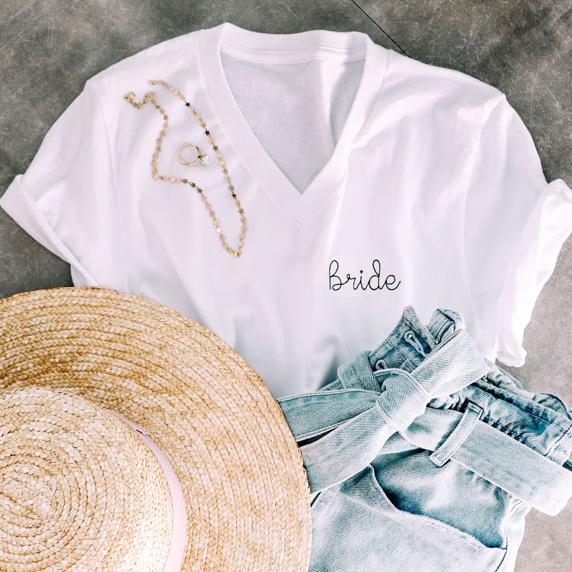 flat lay of v neck t-shirt with bride embroidered in black thread in a stitch font on the left chest paired with a hat, jewelry, and denim shorts