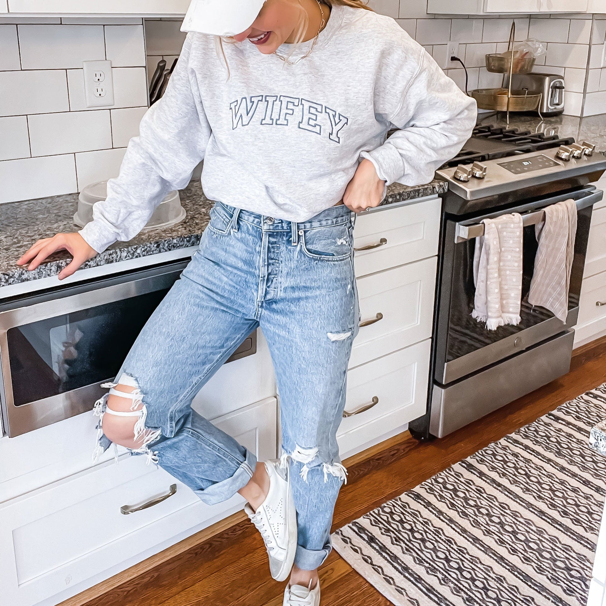 girl posing in her kitchen wearing a crewneck sweatshirt in ash gray with wifey embroidered in a blue thread and athletic block font