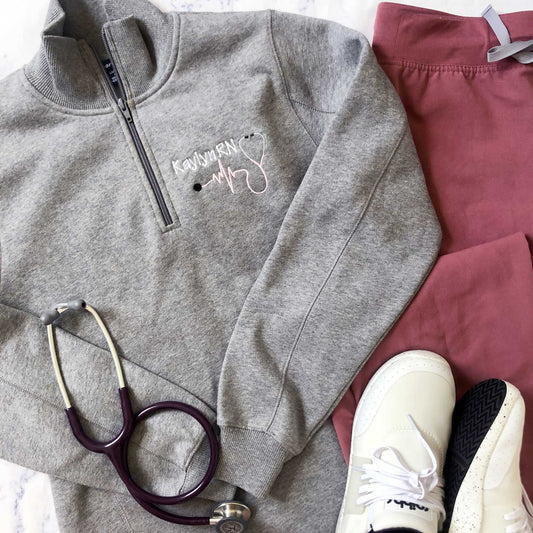 flat lay image of a gray ladies quarter zip with a personalized nurse embroidery design, scrub pants, tennis shoes, and a stethoscope