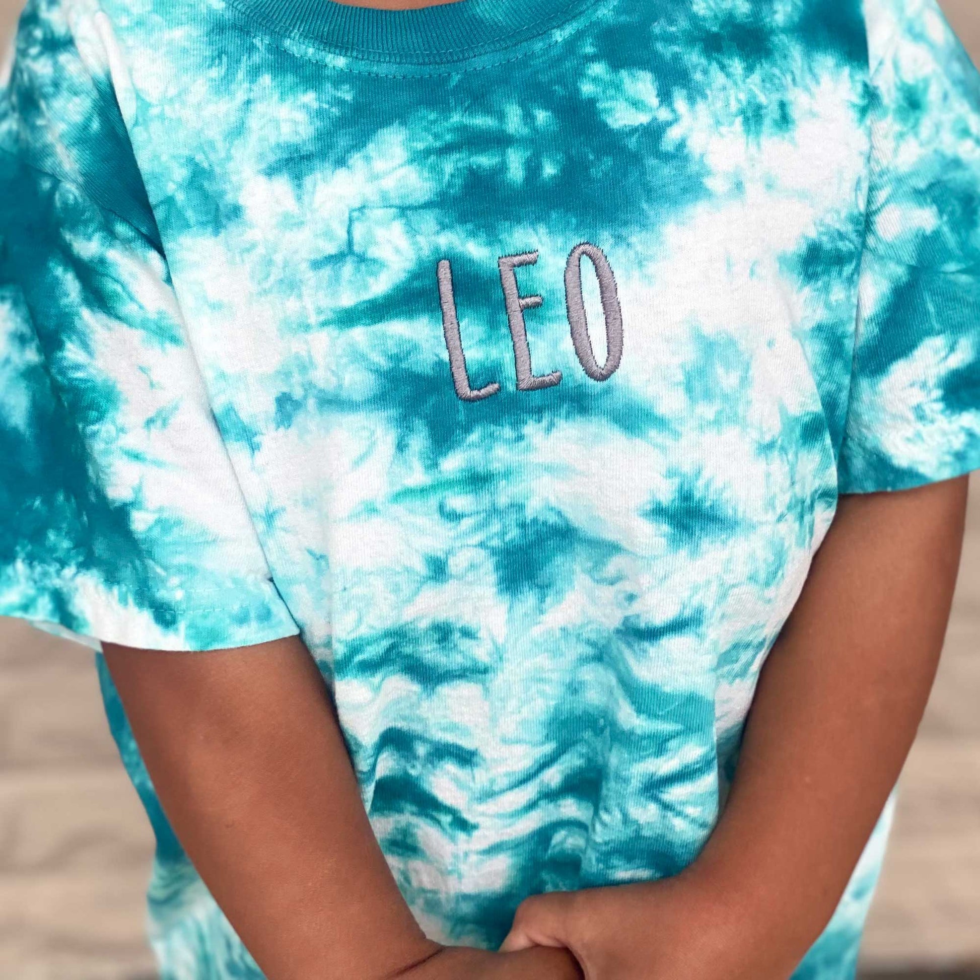 close up photo of a youth crystal tie dye t-shirt in a teal blue and white with a name embroidery in a dark gray thread on the center of the chest 