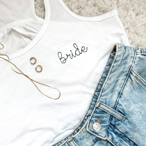 flat lay of white flowy bella and canvas tank top with bride embroidered in black thread paired with jewelry and jean shorts 