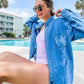 woman sitting by a pool with a faded blue denim button down shirt worn over lavender bathing suit 