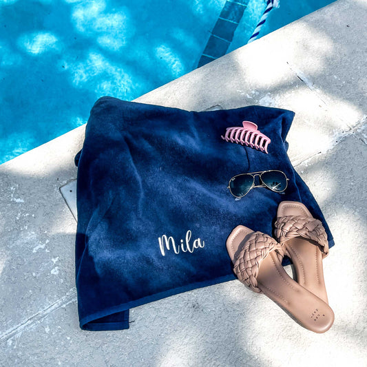 image of a solid navy blue beach towel featuring a full name embroidery along the bottom folded next to the edge of a pool