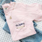 Blossom pocket t-shirt with teacher name and pencil embroidery laying folded over blue jeans on white quilted blanket