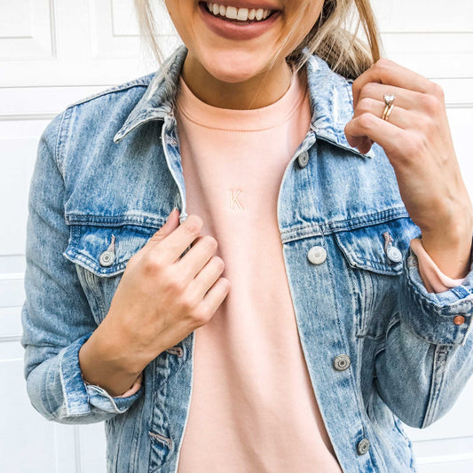 girl wearing a jean jacket over light peach crewneck sweatshirt with mini initial embroidered on the center chest