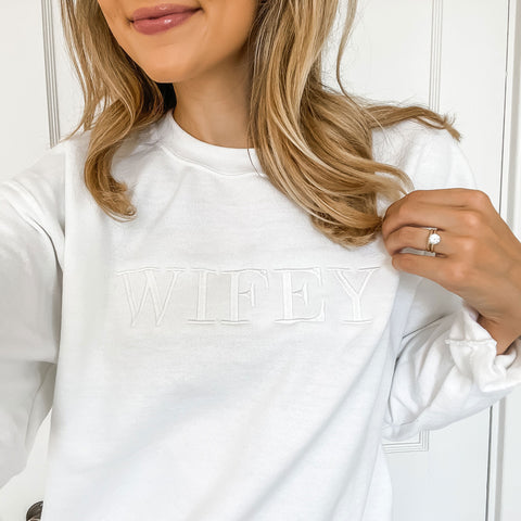 woman wearing a white crewneck sweatshirt with wifey embroidered in serif block font across the chest