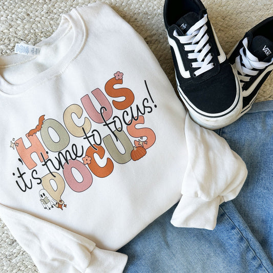 flat lay image of an outfit featuring jeans, black vans, and a crewneck sweatshirt with a colorful halloween teacher print reading hocus pocus it's time to focus