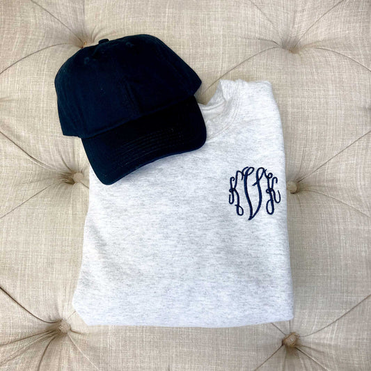 ash gray sweatshirt with custom personalized monogram embroidery in navy thread on the left chest folded on top of an ottoman next to a navy baseball cap