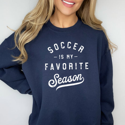 woman wearing a navy pullover crewneck sweatshirt with a printed dtg distressed design across the front reading 'soccer is my favorite season' in white ink