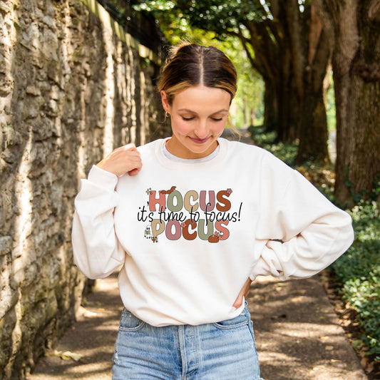 woman wearing a crewneck sweatshirt with a hocus pocus themed printed design across the chest reading hocus pocus it's time to focus!