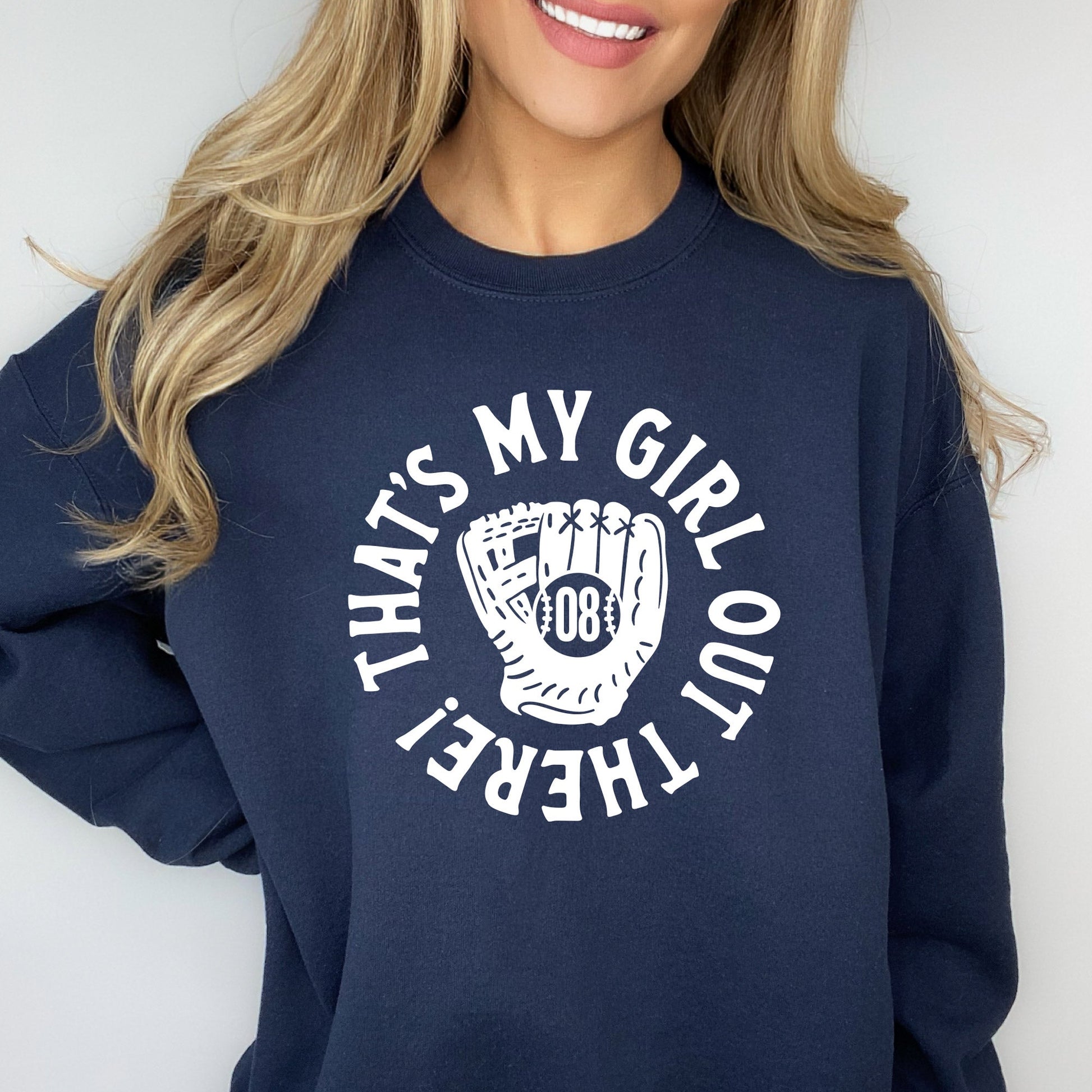 woman wearing a navy blue crewneck sweatshirt with a printed design of a baseball mitt with player number in the center and 'that's my girl out there!' in a circle around it.