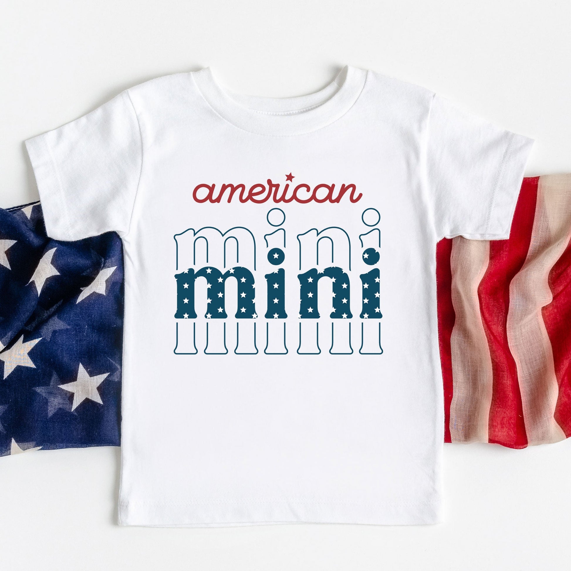 white youth tee laying on an american flag with an 'american mini' printed design in red and blue