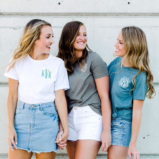 light blonde girl wearing a white crewneck tee with blue personalized monogram embroidery, brunette wearing a deep heather gray tee with custom three letter embroidery, and dark blonde wearing a blue shirt with script monogrammed embroidery