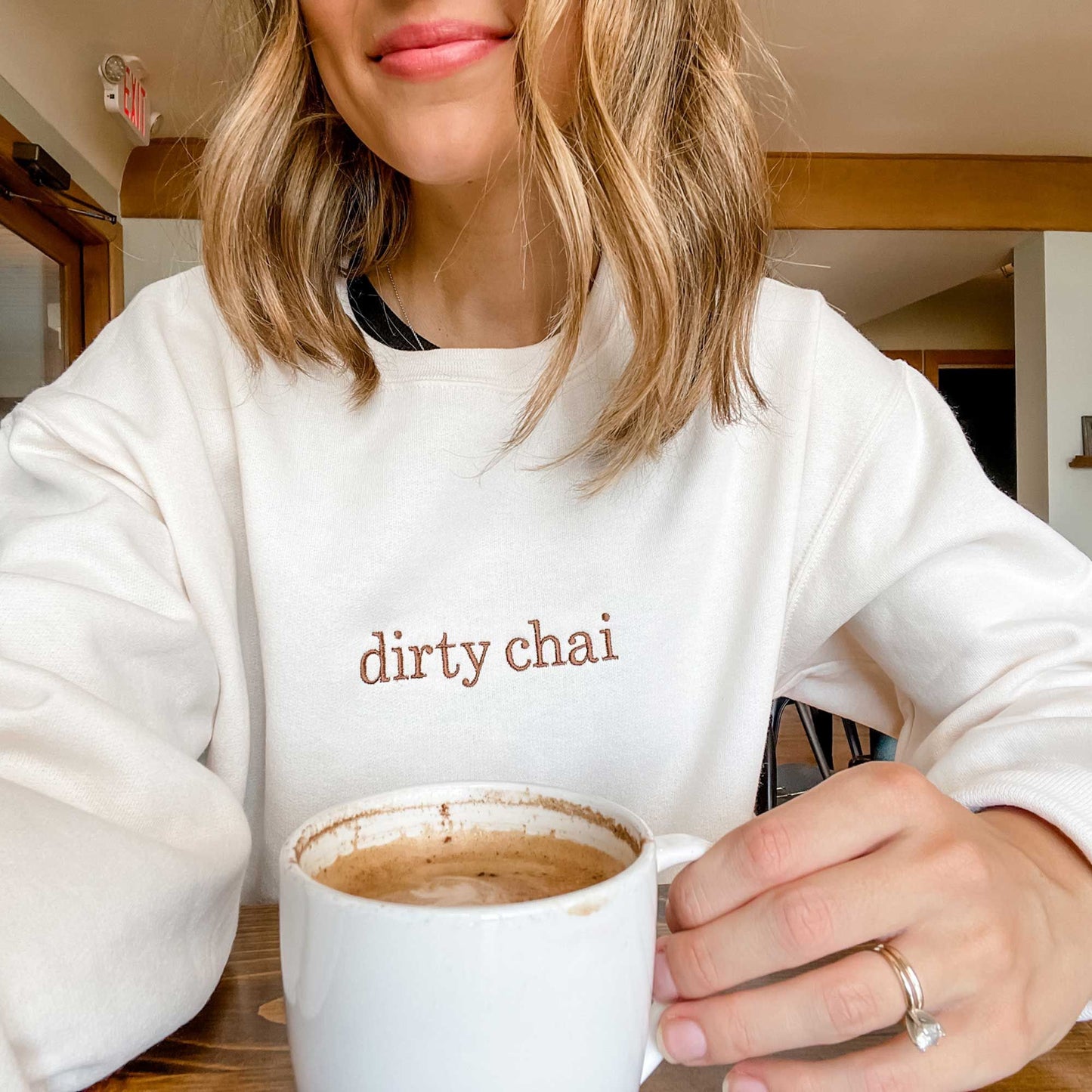 woman wearing a crewneck cream colored sweatshirt with her custom coffee order embroidered across the center chest