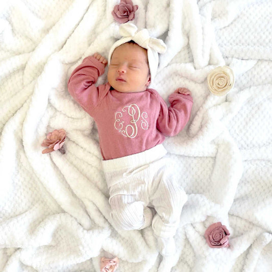 newborn baby girl laying on a blanket wearing a headband, mauve pink long sleeve bodysuit featuring a script three letter monogram and leggings