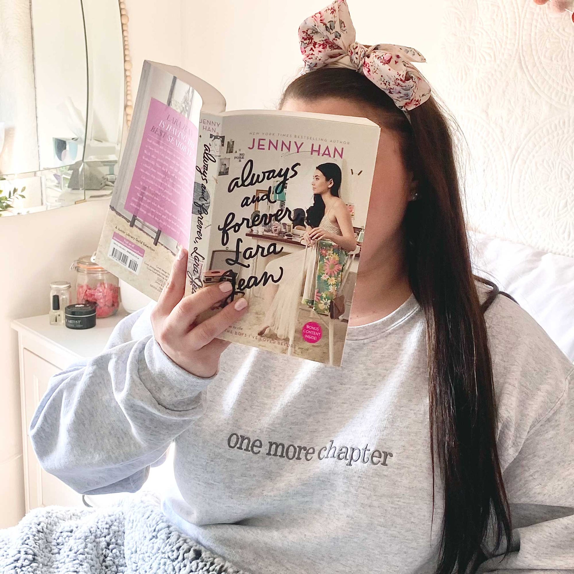 girl sitting in bed and reading a book wearing a cotton pullover sweatshirt with one more chapter embroidered across the chest