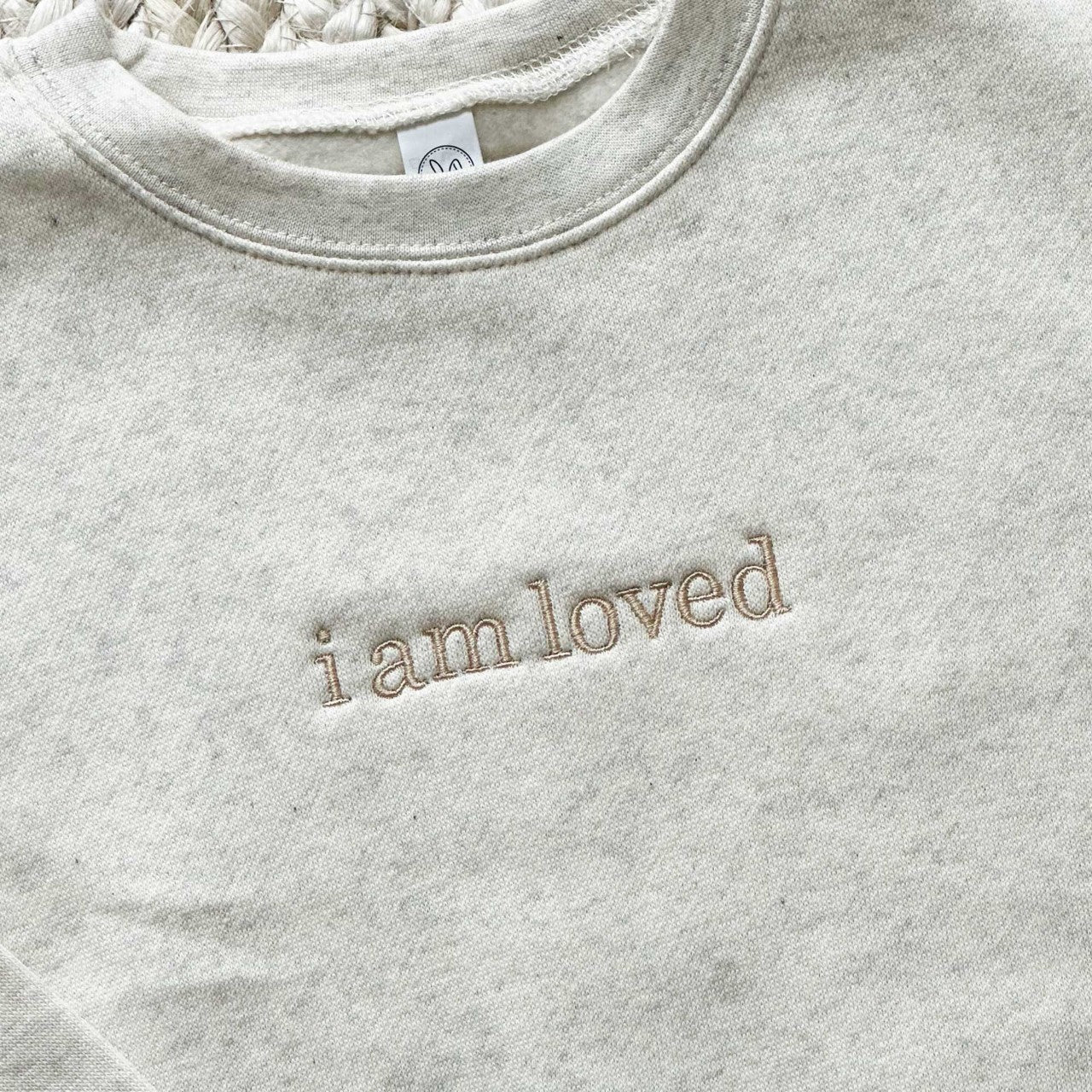 close up of the embroidery i am loved on a heather oatmeal crewneck sweatshirt